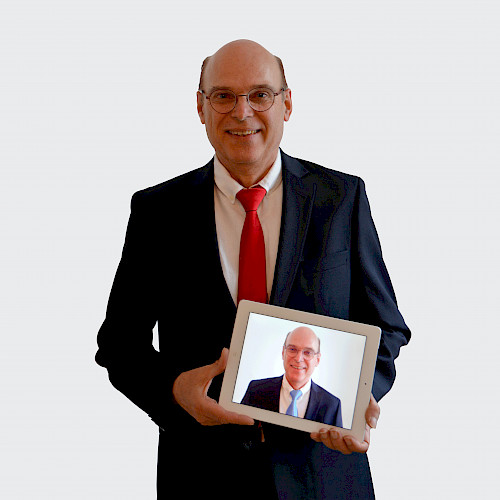 Porträt von Herr Clemens, Steuerberater in dem Steuerbüro Clemens steht lächelnd im Anzug mit eine Bild von sich in der Hand.