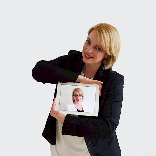 Porträt von Frau Clemens, Steuerberaterin in dem Steuerbüro Clemens steht lächelnd im Anzug mit eine Bild von sich in der Hand.